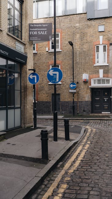 A narrow street scene in an urban area showing multiple traffic signs attached to black metal posts, including directional arrows indicating left and right turns, situated on a section of cobblestone